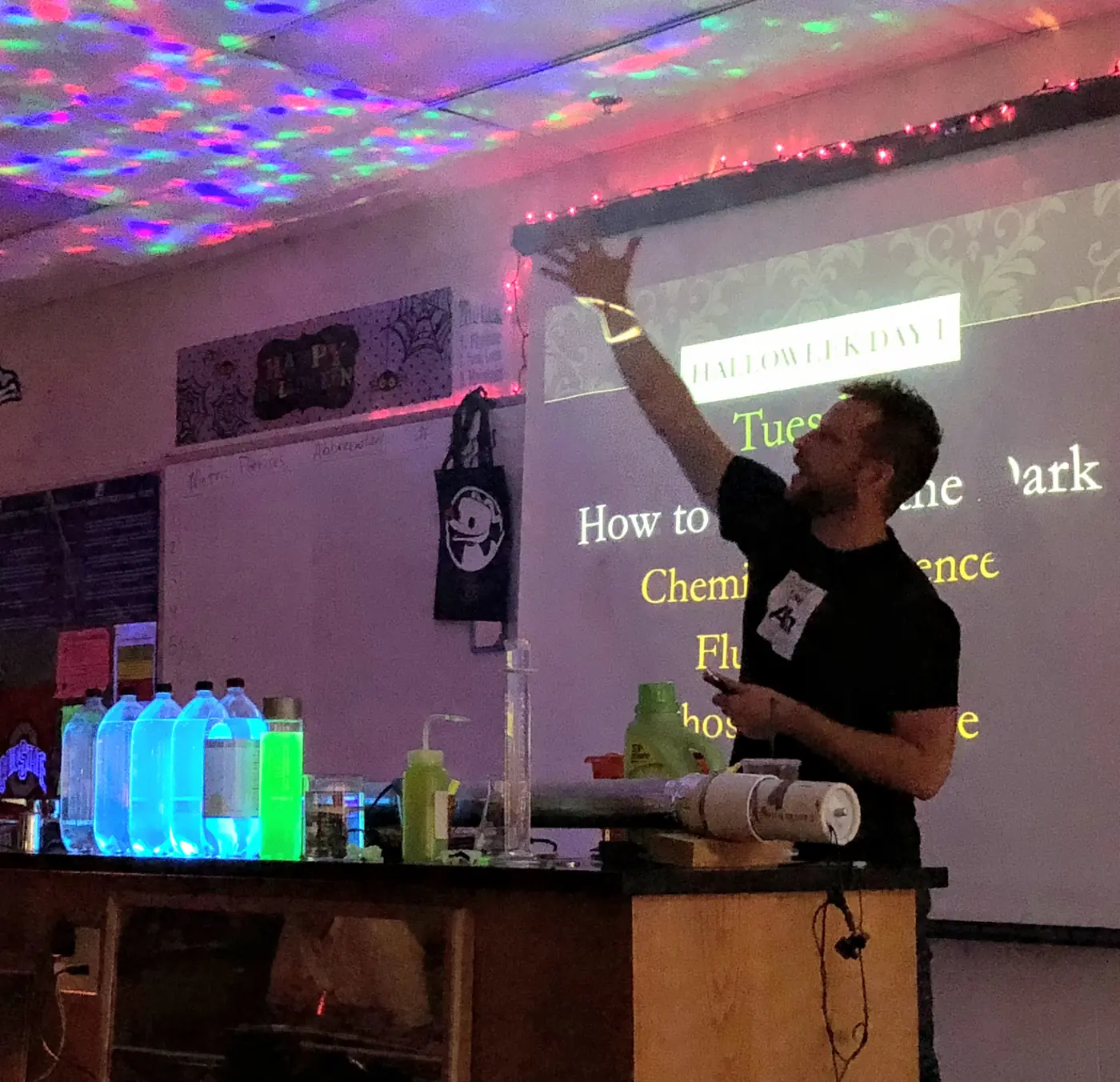 Hutch reaching toward a projected slide about chemistry in the dark, with glowing bottles on the lab bench